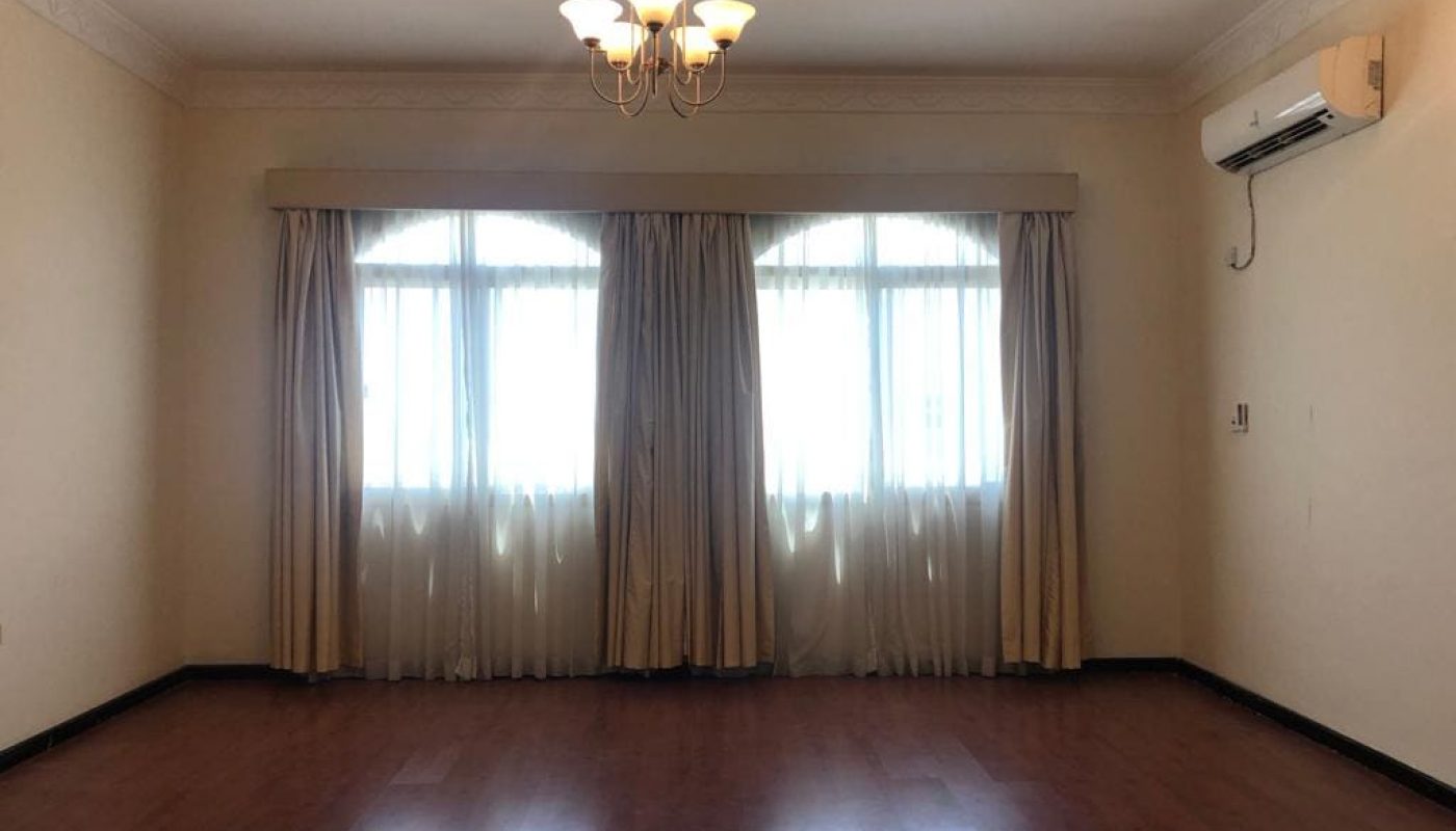 An empty semi-furnished room with wooden flooring, beige walls, two covered windows, and an air conditioner mounted on the right wall. A ceiling light fixture is centrally positioned in this spacious villa with 5 bedrooms.