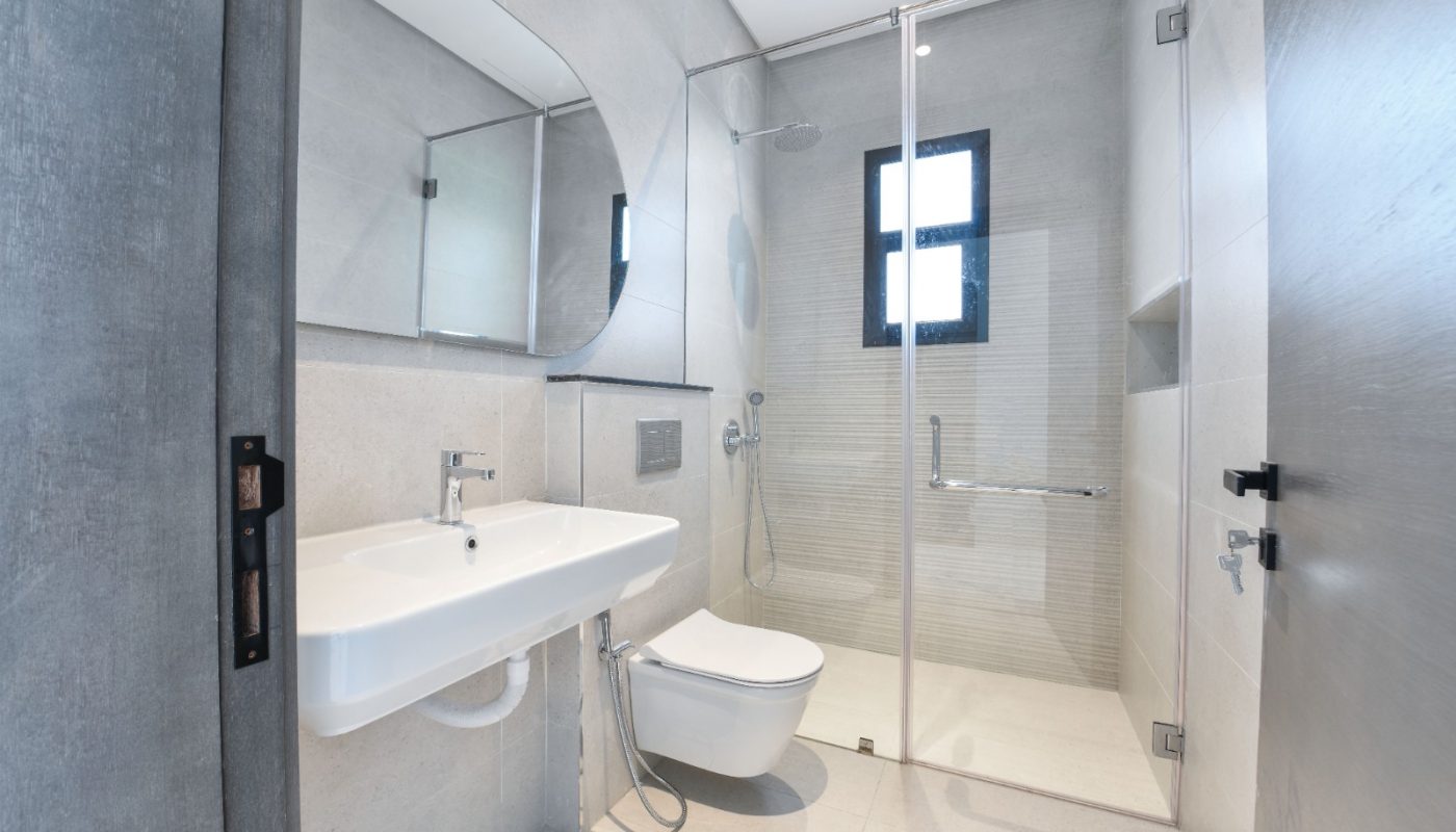 Modern bathroom with wall-mounted sink and toilet, glass-enclosed shower, large mirror, and a small window. Surfaces feature light-colored tiles and minimalist fixtures.