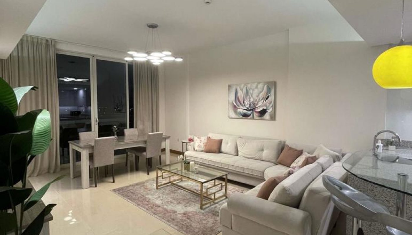 Modern living room with beige sofas, dining table, potted plant, bar stools, and artwork on the wall, illuminated by overhead lighting.