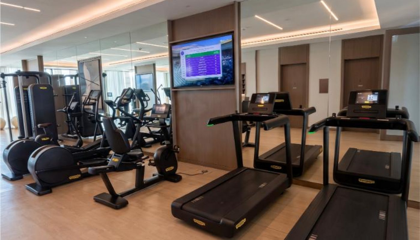 Modern gym room with treadmills, elliptical machines, exercise bikes, a weight machine, wall mirrors, wood flooring, and a wall-mounted TV displaying workout data.