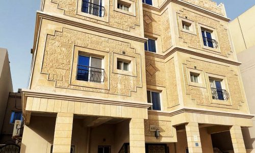 Modern multi-story residential building for sale with textured beige facade, balconies, and parked cars underneath in the New Hidd Area.