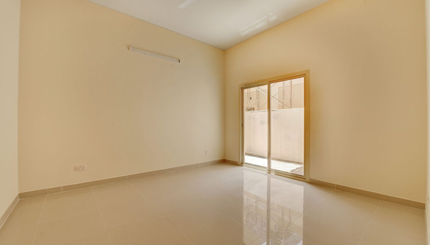 An empty room with beige tiled floor, cream walls, a ceiling fan, and a glass sliding door leading to a small patio or balcony.