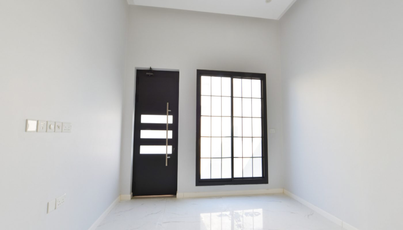 A small, empty room with white walls, a black door, a large window with grid panes, and a white tiled floor.