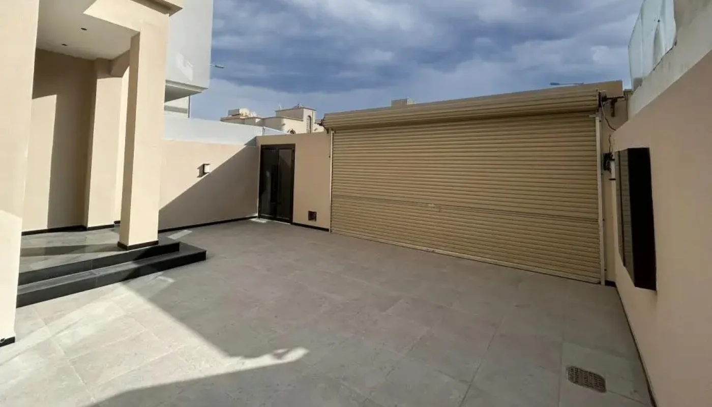 A paved courtyard with beige walls, a closed roll-up garage door, black steps leading to an entrance, and a partly cloudy sky above.