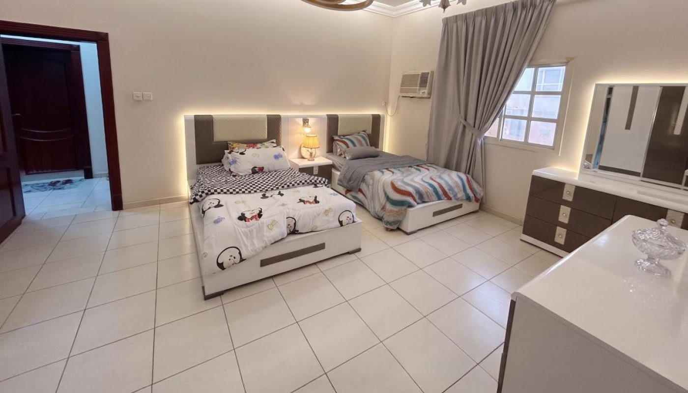 A tidy bedroom with two single beds, modern white furniture, tiled floor, a window with gray curtains, and soft ambient lighting.