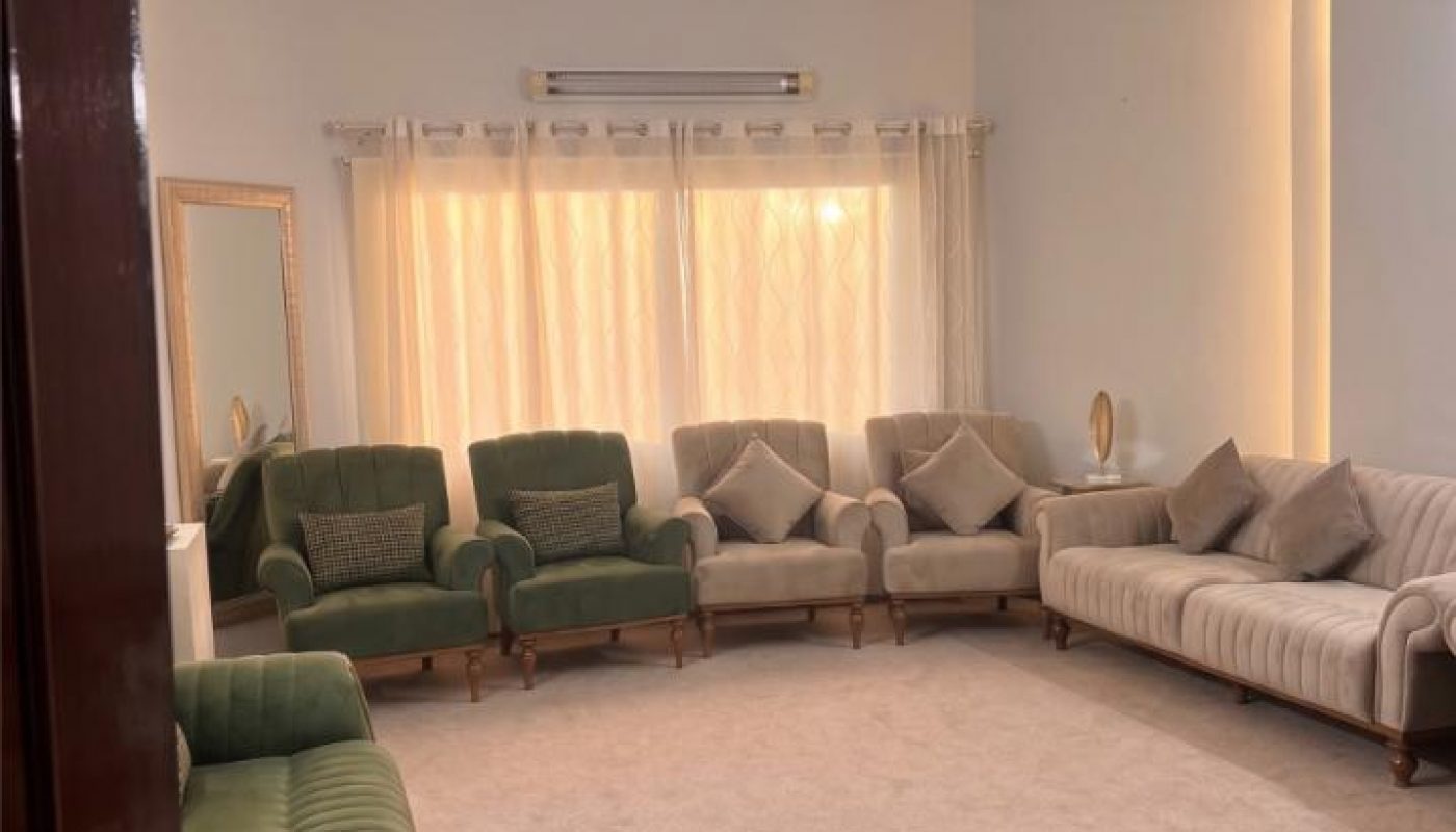 A living room with green and beige sofas, a beige carpet, a ceiling fan, a large window with curtains, and a wall-mounted air conditioner.