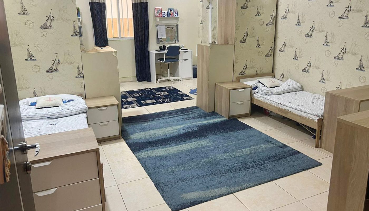 A tidy bedroom with two single beds, a blue rug, desks, bookshelves, and patterned wallpaper featuring sketches of the Eiffel Tower.