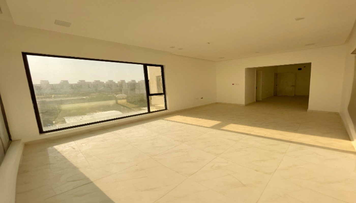An empty, unfurnished room with large windows, tiled floor, white walls, and sunlight streaming in. A cityscape is visible through the window.