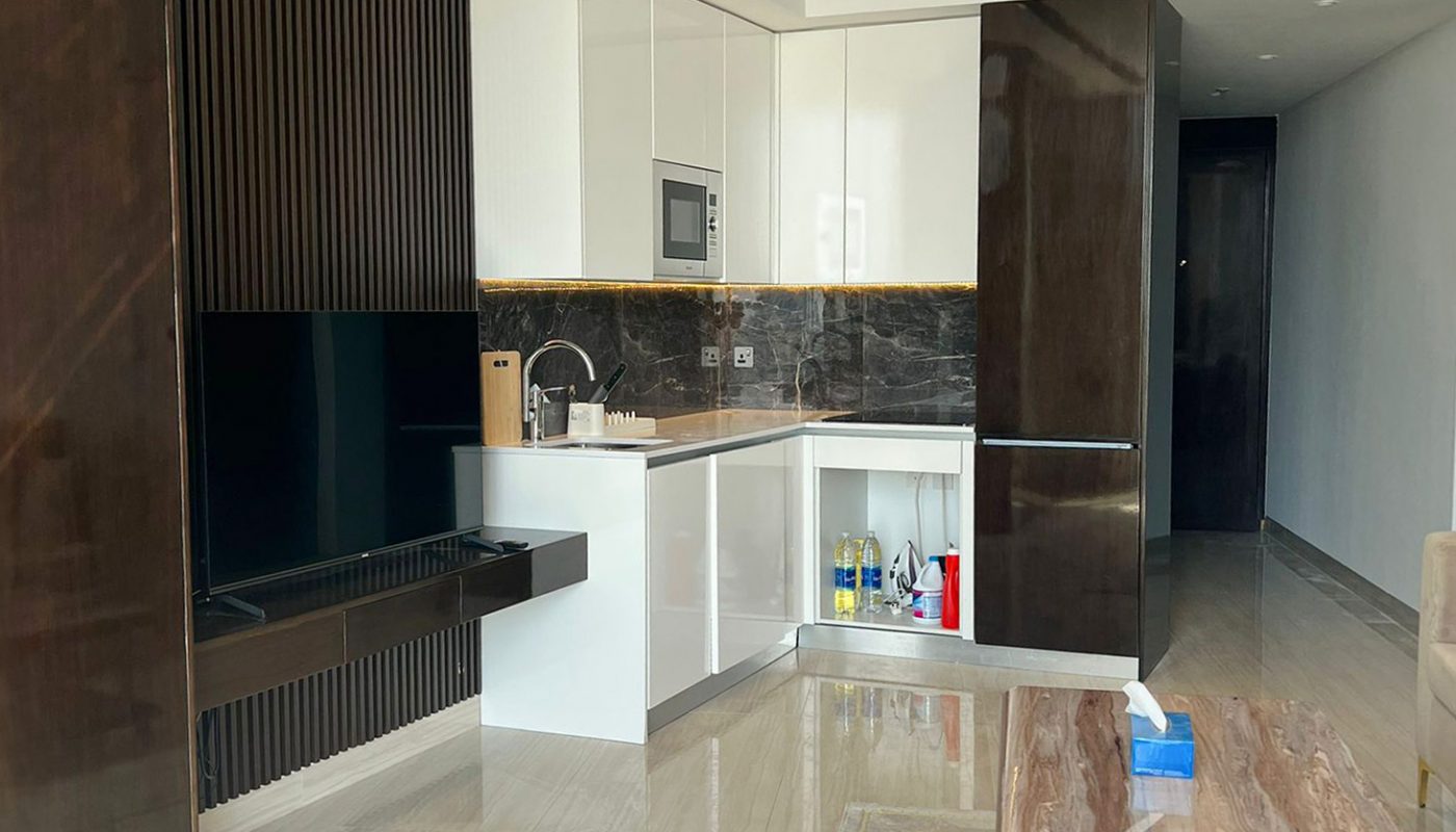 Modern studio apartment interior with a small kitchenette, dark wood accents, a mounted TV, a marble coffee table, and a bed in the foreground.