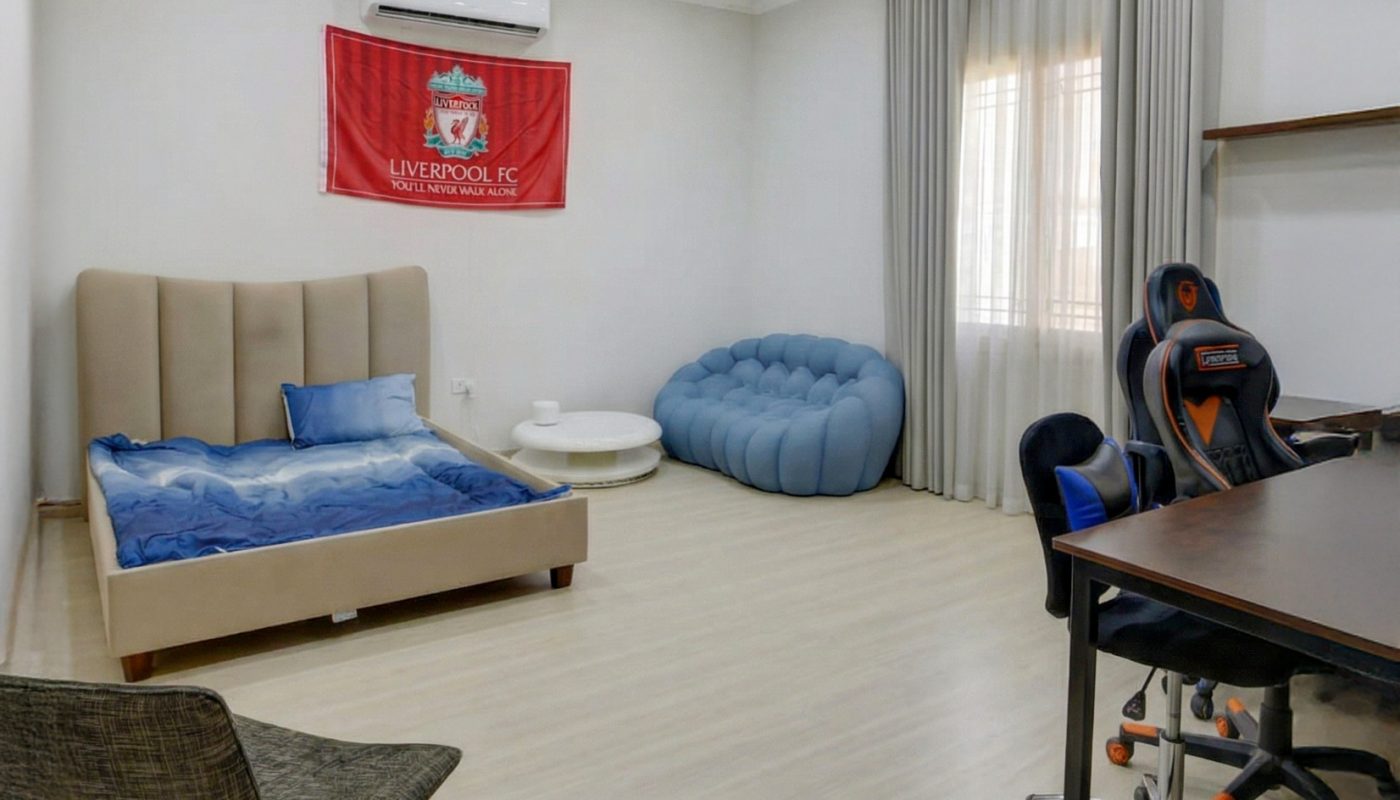 A modern bedroom with a bed, blue sofa, desk and gaming chair, wall-mounted shelves, a gray chair, and a Liverpool FC flag on the wall.