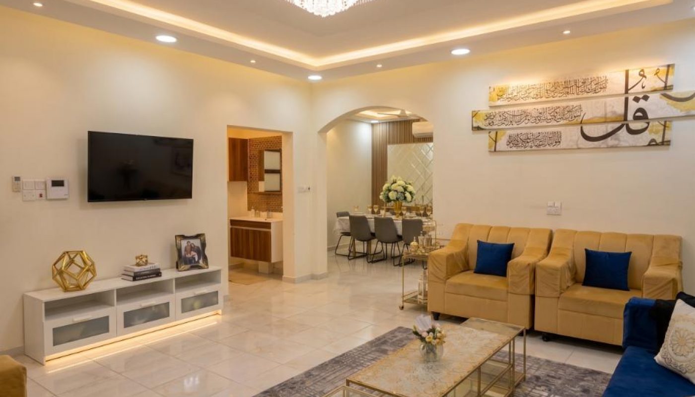 A modern living room with beige sofas, blue cushions, a wall-mounted TV, a glass coffee table, and a chandelier, leading to a dining area in the background.