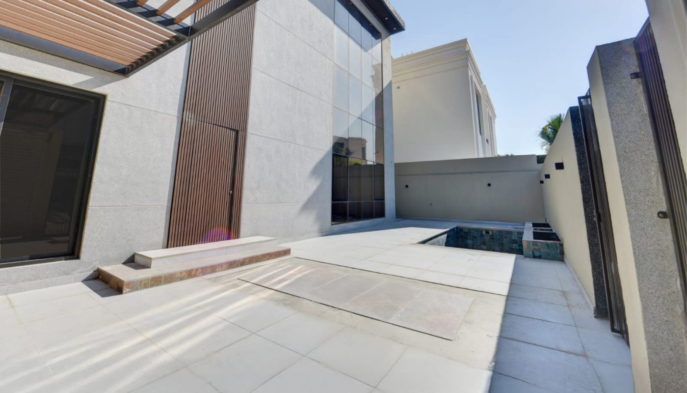 Modern minimalist courtyard with white tile flooring, a small pool, wooden pergola, and tall glass windows; sunlight visible in the corner of the image.