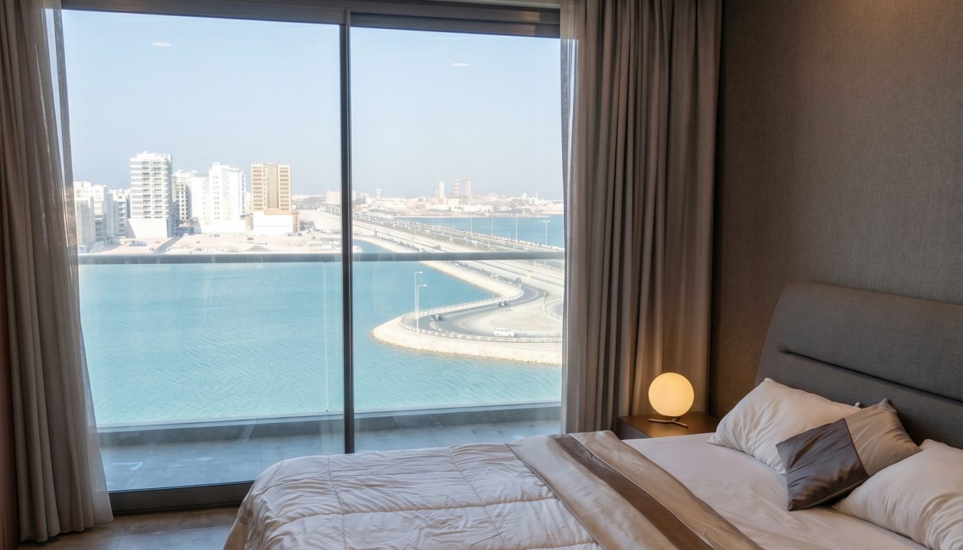 A modern bedroom with a large bed and floor-to-ceiling window overlooks a city skyline and waterfront under a clear sky.