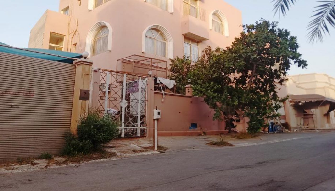 A three-story pink building with arched windows and a metal gate faces a quiet, empty street under a clear sky, with trees and plants nearby.