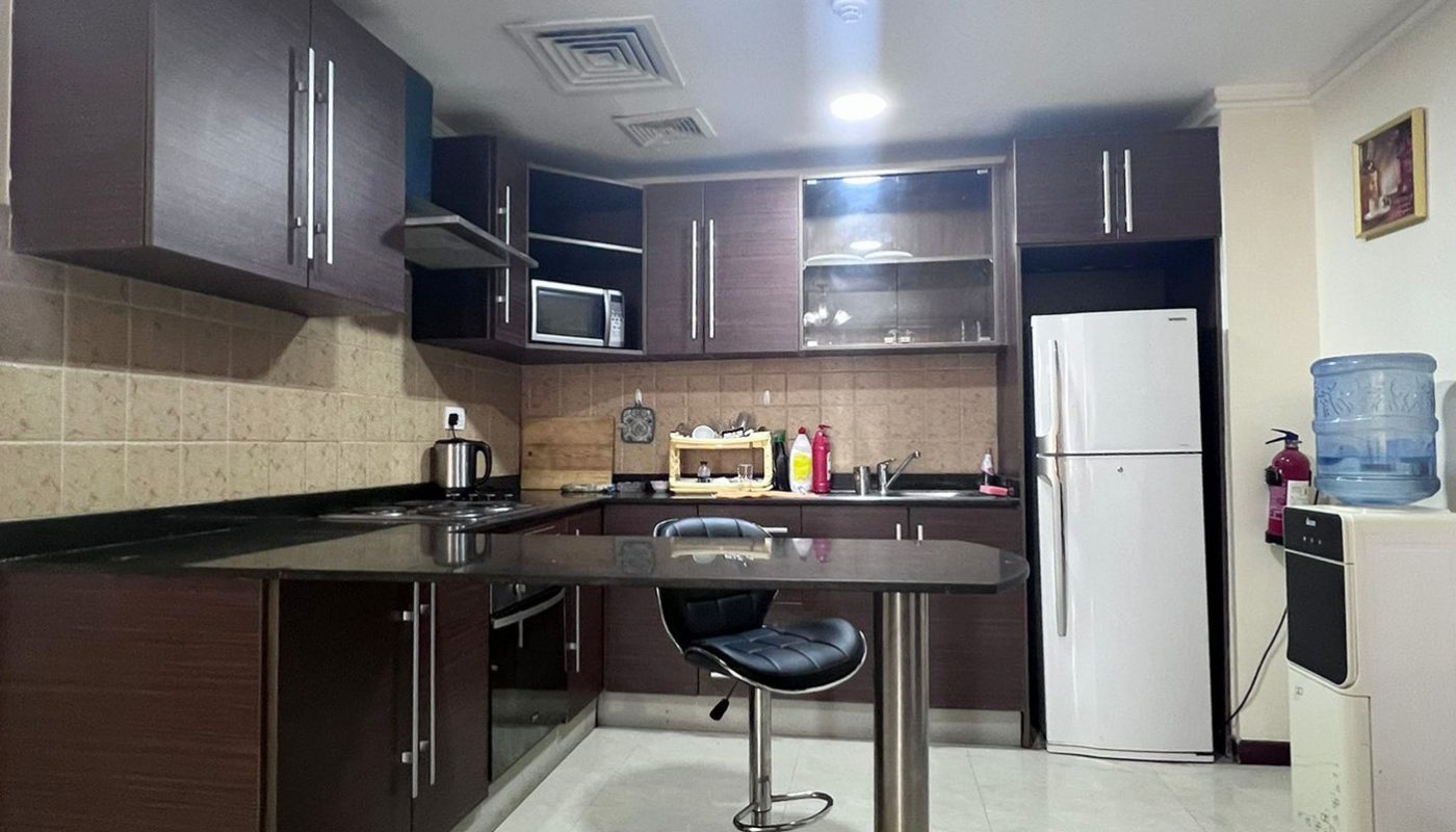 Modern kitchen with dark wood cabinets, a central island with a stool, refrigerator, microwave, water dispenser, and countertop appliances under bright ceiling lights.