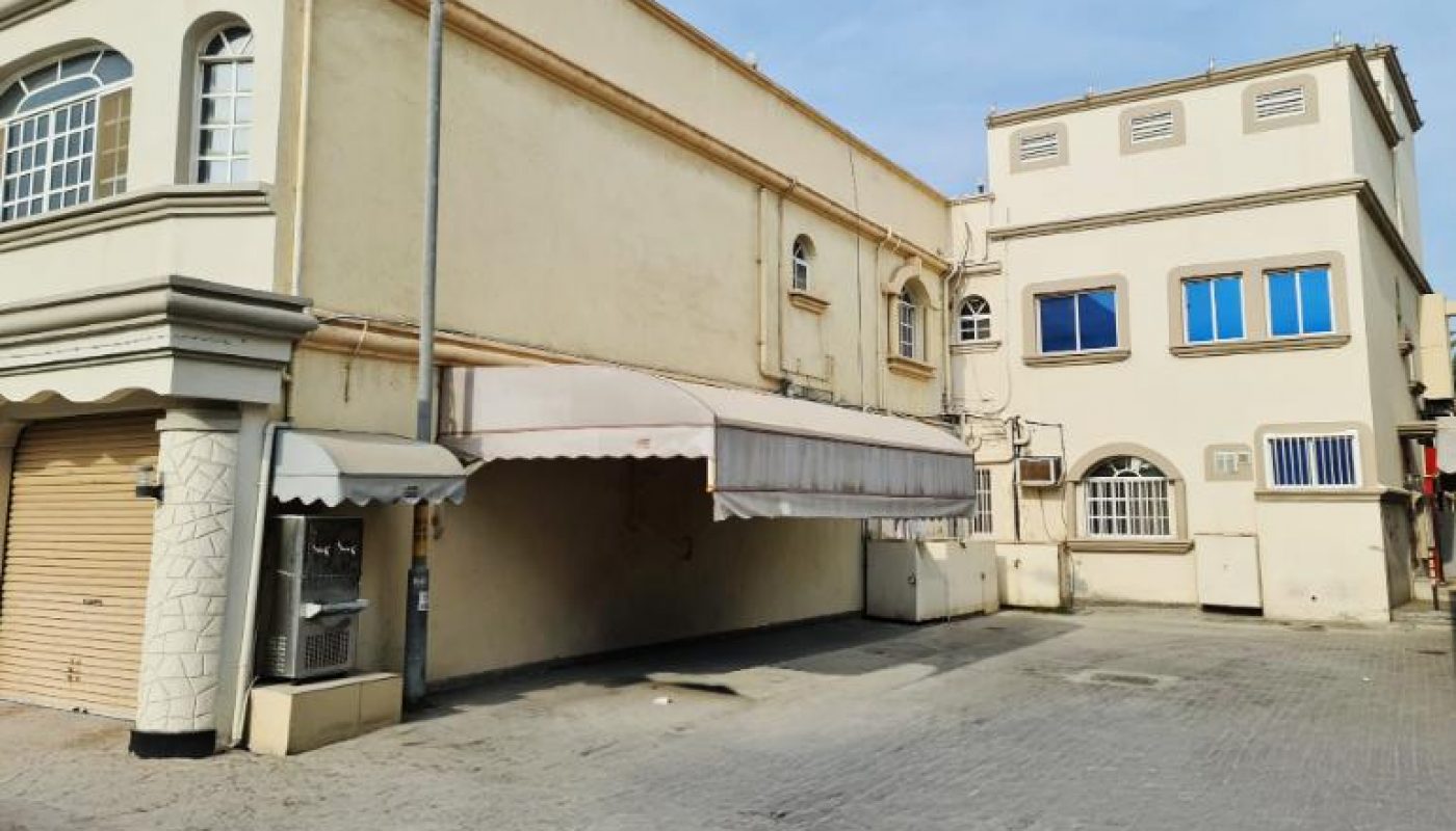 Two beige buildings with blue and white windows are shown beside a paved lot under a blue sky. One building has an awning and a closed garage door.