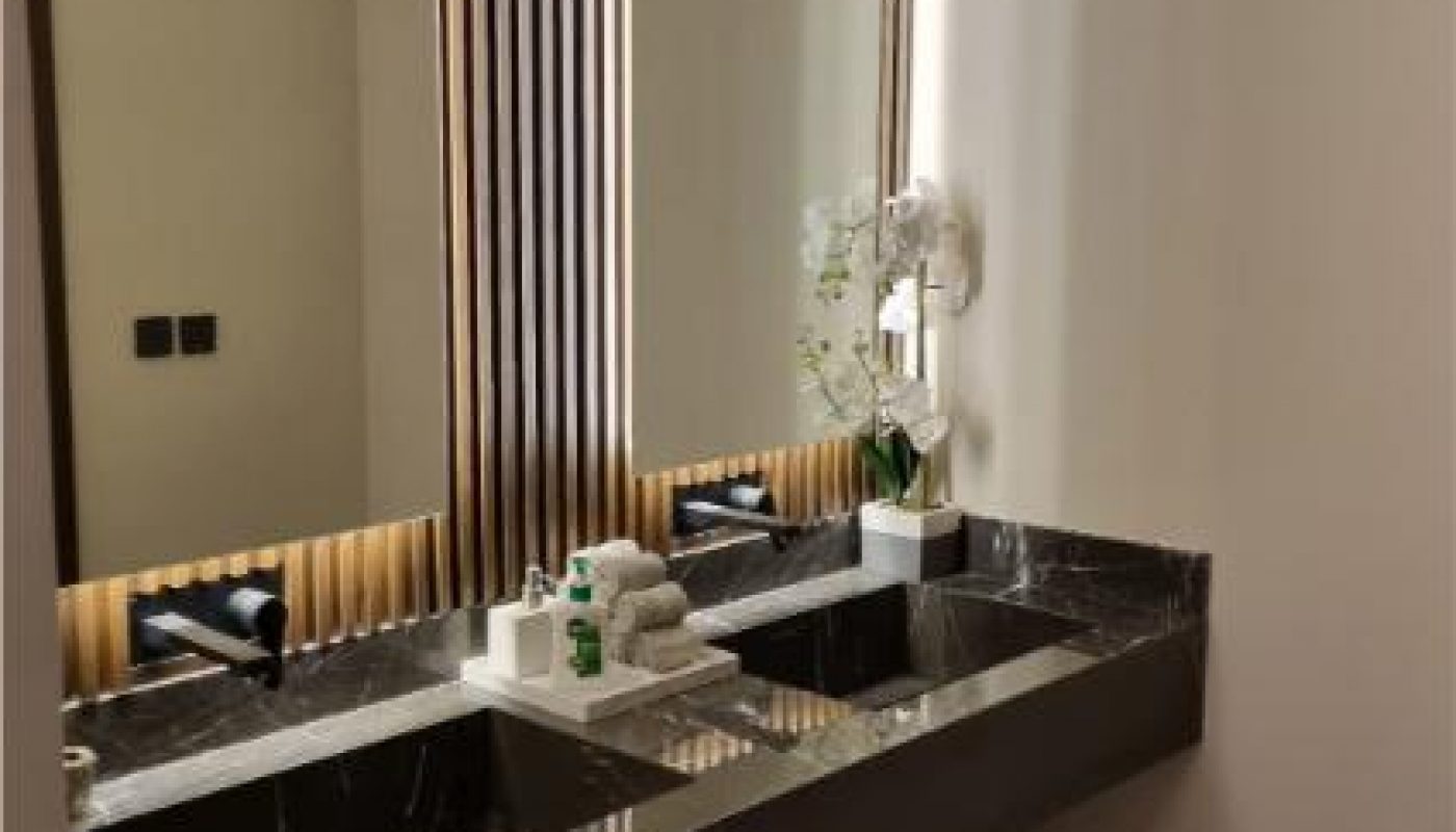 Modern bathroom with two black marble sinks, wooden slat wall, large mirrors, and a countertop holding towels, toiletries, and a white potted plant.