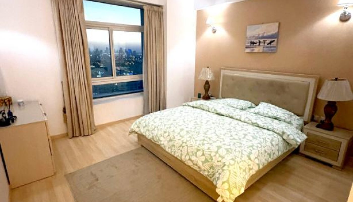 A neatly made bed with a green patterned cover sits in a modern bedroom with light wood floors, beige walls, two lamps, a dresser, and a window with beige curtains showing a city view.