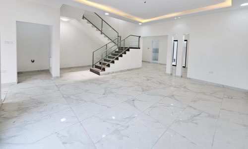 A modern, spacious room in a luxurious villa with white marble flooring, a staircase with a glass railing, recessed lighting in the ceiling, and white walls. Two open doorways lead to adjacent rooms in this stunning property for sale.