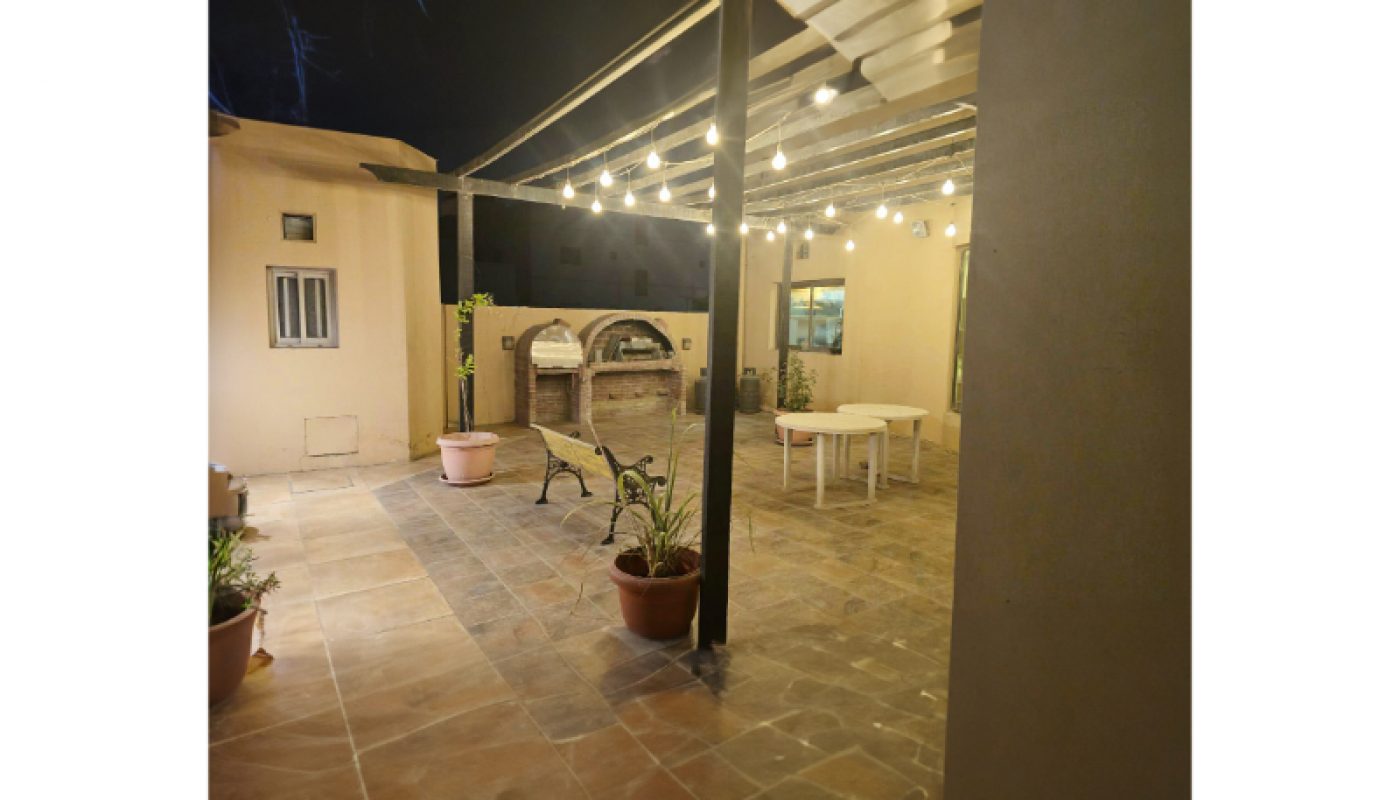 Covered patio at night with string lights, potted plants, a table, chairs, and an outdoor oven against the far wall.