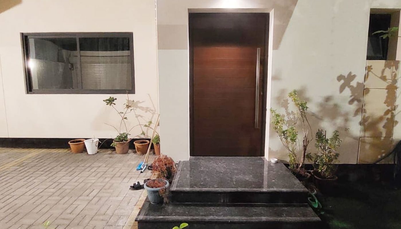 A house entrance at night, featuring a lit doorway with dark wooden door, steps, potted plants, and a window on the left.