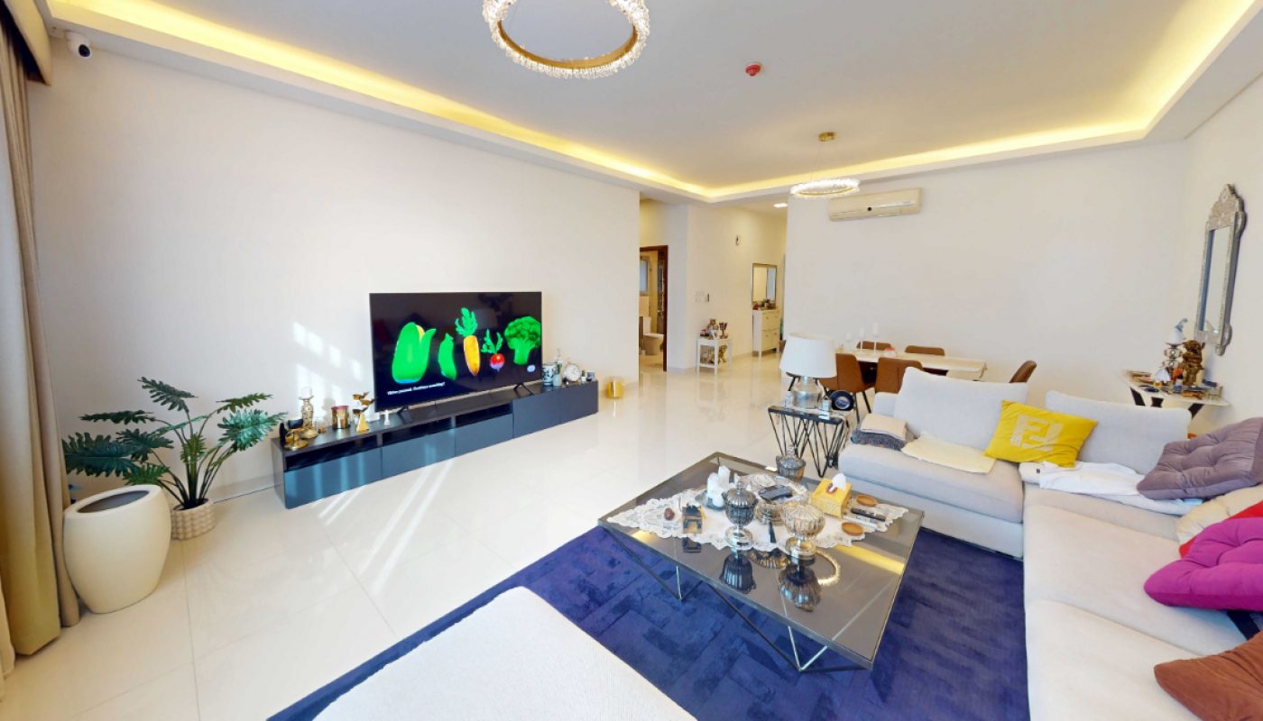 Modern living room with white walls, a sectional sofa with colorful pillows, a glass coffee table, a flat-screen TV, and decorative lighting.