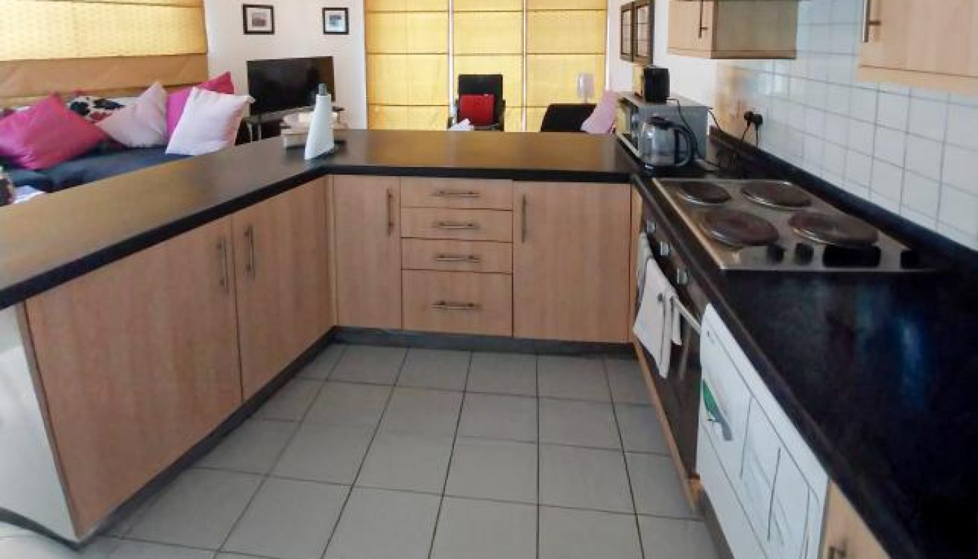 Modern kitchen with light wood cabinets, black countertops, stove, oven, and small appliances; living area with colorful pillows visible in the background.