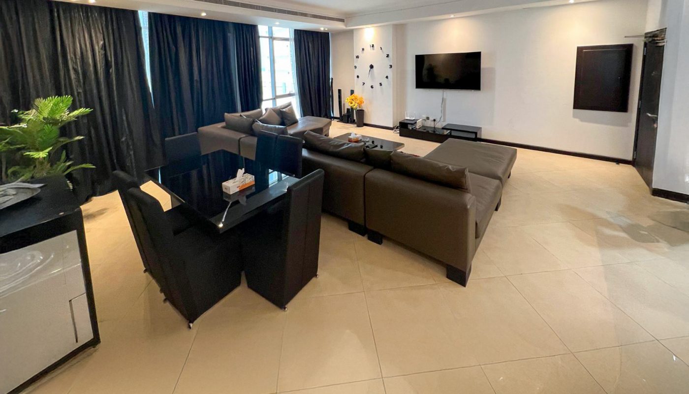 Spacious modern living room with brown sofas, a glass dining table, wall-mounted TV, and large windows with dark curtains.