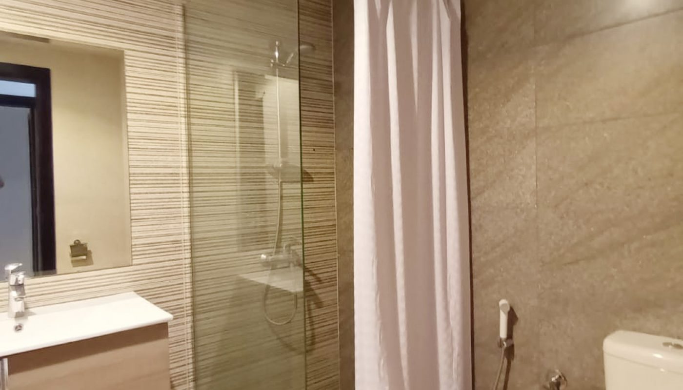 Modern bathroom with a glass-enclosed shower, white curtain, wall-mounted sink, toilet, and handheld bidet on gray tiled walls and floor.