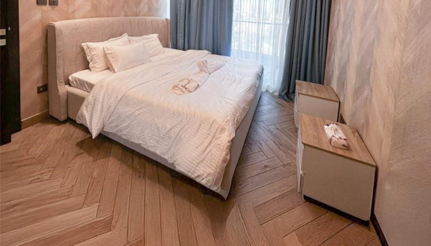 Modern bedroom with a double bed, white bedding, two bedside tables, wooden chevron-pattern floor, and floor-to-ceiling window with sheer and dark curtains.