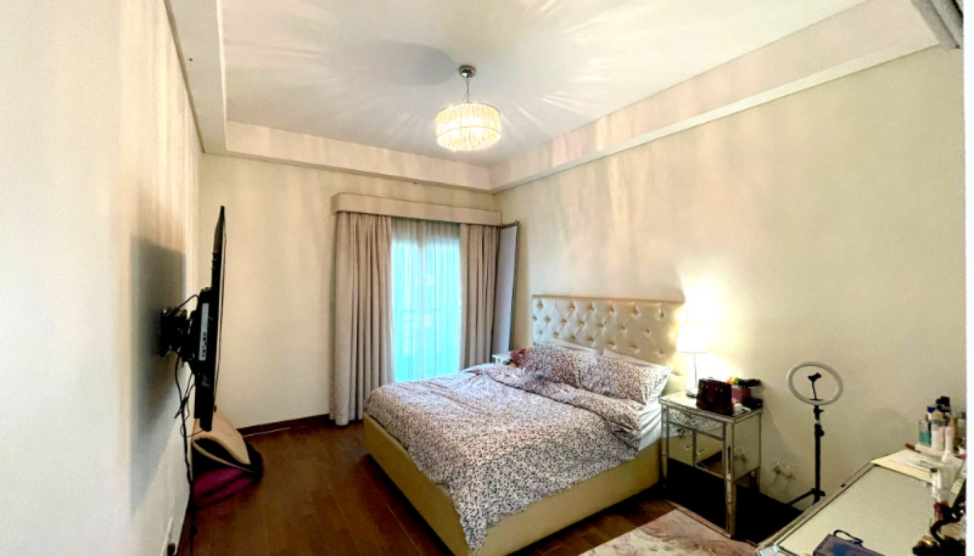 A neatly made bed with a tufted headboard sits against the wall in a bedroom with wooden floors, a TV mounted opposite, a dressing table, a rug, and a window with floor-length curtains.