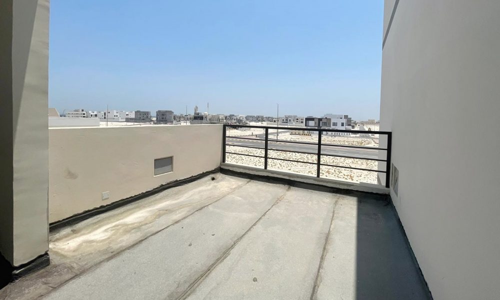 View of a balcony in an elegant villa with concrete flooring, partial shade, and metal railings overlooking a bright, distant cityscape under a clear blue sky. This stunning property in Diyar Al Muharraq is now for sale.