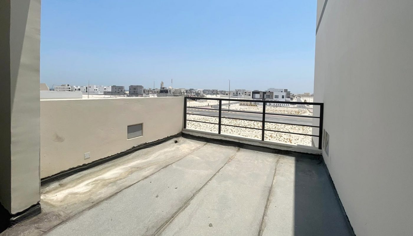 View of a balcony in an elegant villa with concrete flooring, partial shade, and metal railings overlooking a bright, distant cityscape under a clear blue sky. This stunning property in Diyar Al Muharraq is now for sale.