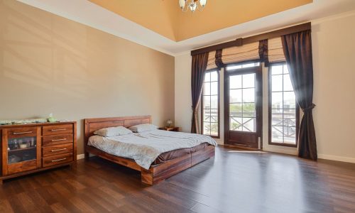 A spacious bedroom with a high ceiling, wooden floor, a bed with white and brown bedding, a wooden dresser, and large windows with curtains allowing natural light to fill the room. This serene space is part of the 3BR Villa in Al Areen, complete with a big garden for outdoor relaxation.