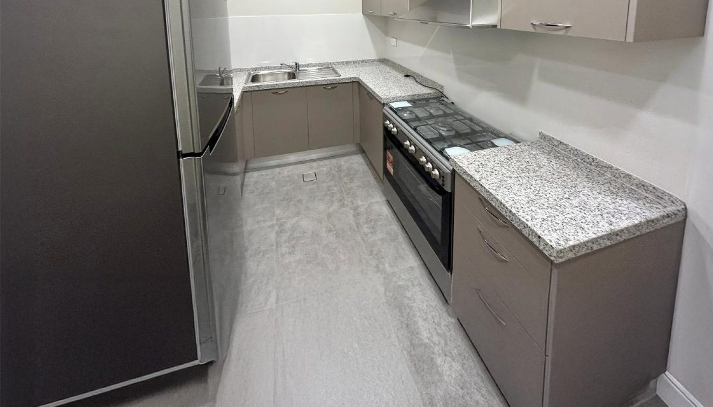 Modern kitchen with grey cabinets, granite countertops, stainless steel fridge, gas stove, sink, and bright overhead lighting.