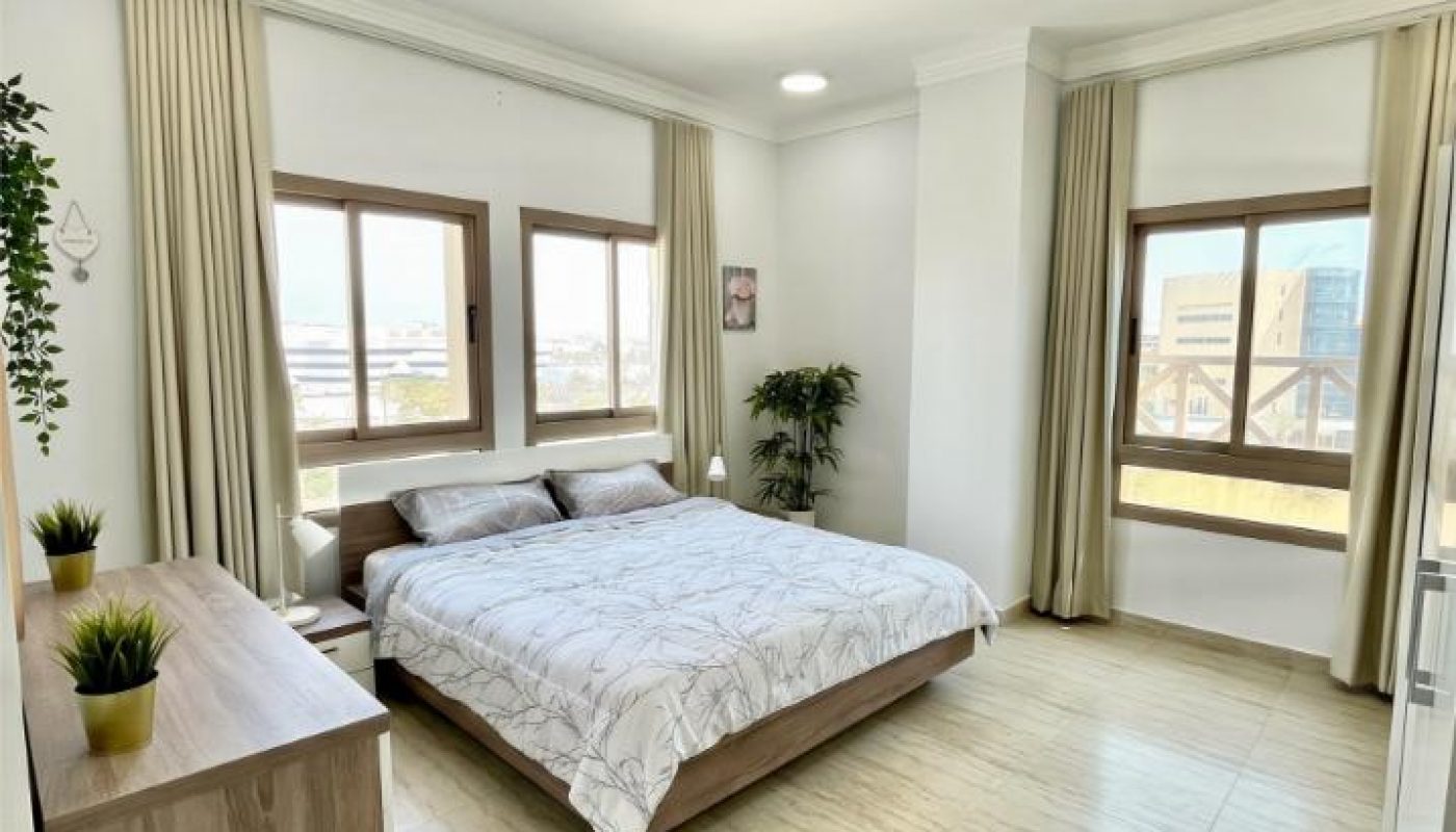 Bright bedroom with large windows, a double bed, light-colored curtains, tile flooring, a wooden dresser with plants, and minimal decor.