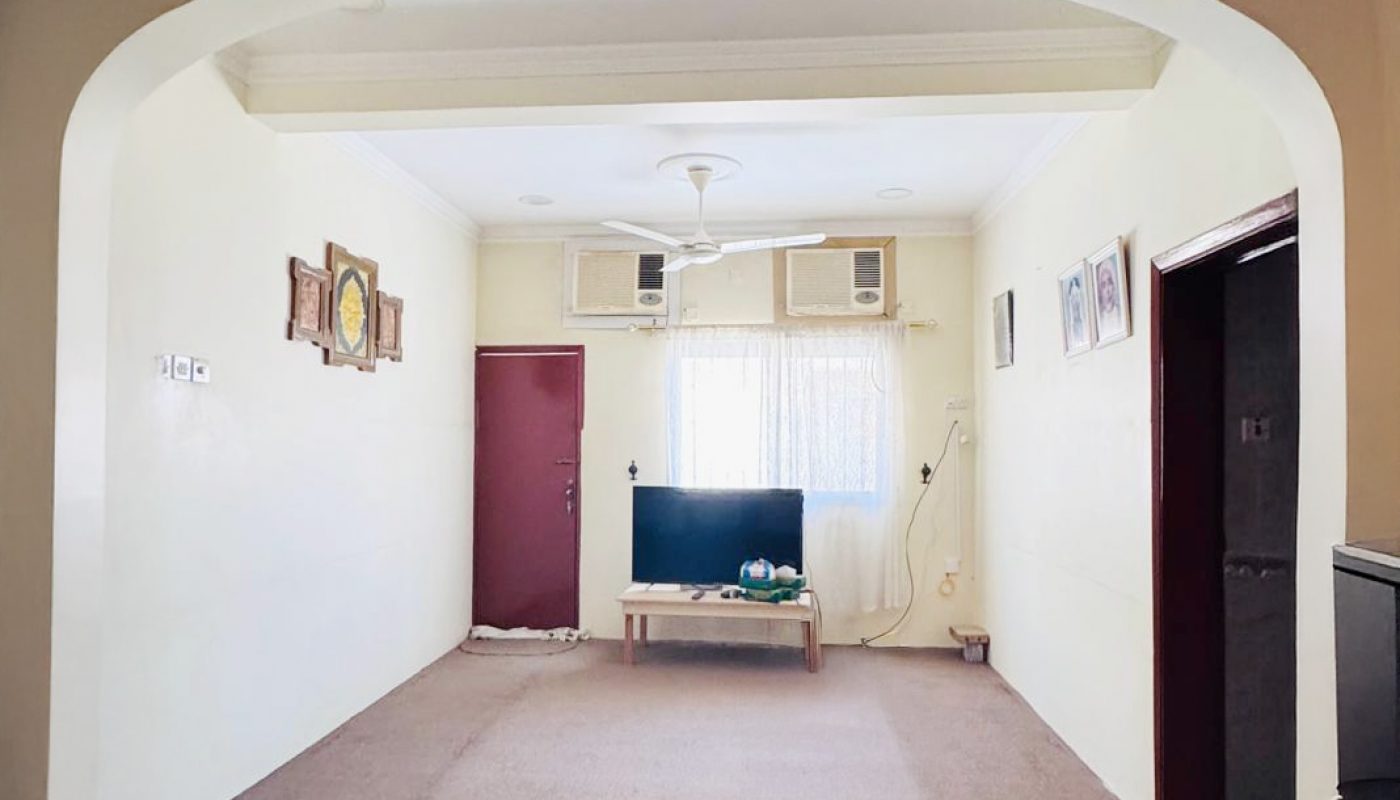 A simple living room with a wall-mounted TV, brown carpet, air conditioners above the window, and framed pictures on cream-colored walls.