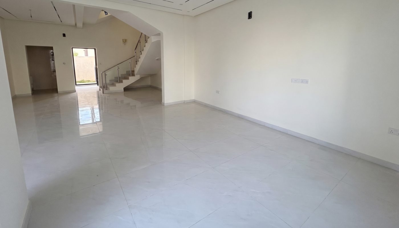 Empty, unfurnished room with white tile flooring, cream-colored walls, ceiling light fixtures, and a staircase leading to an upper floor. Natural light enters from a door and window.