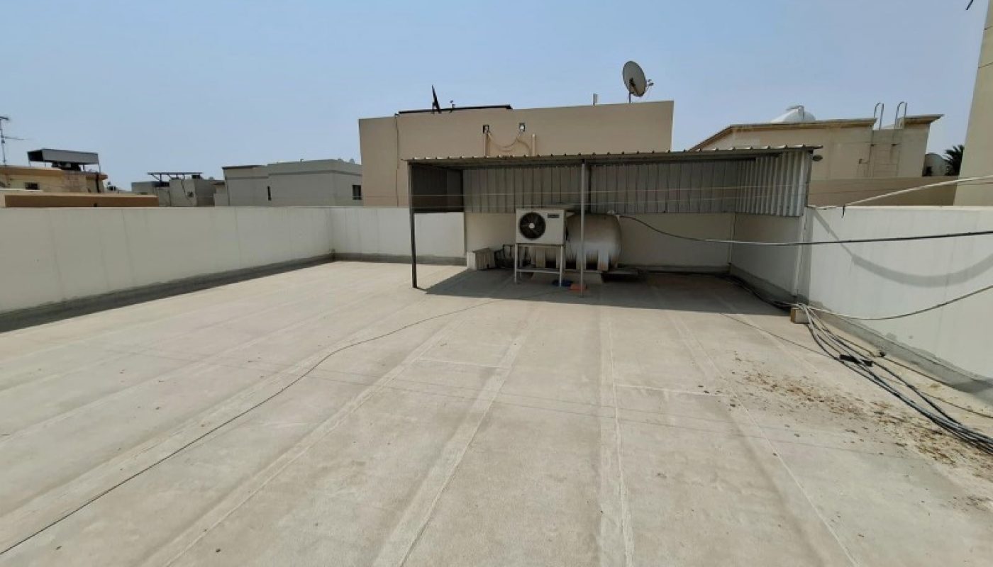 A flat, empty rooftop with a covered structure housing tanks and equipment, surrounded by a white boundary wall under a clear sky.