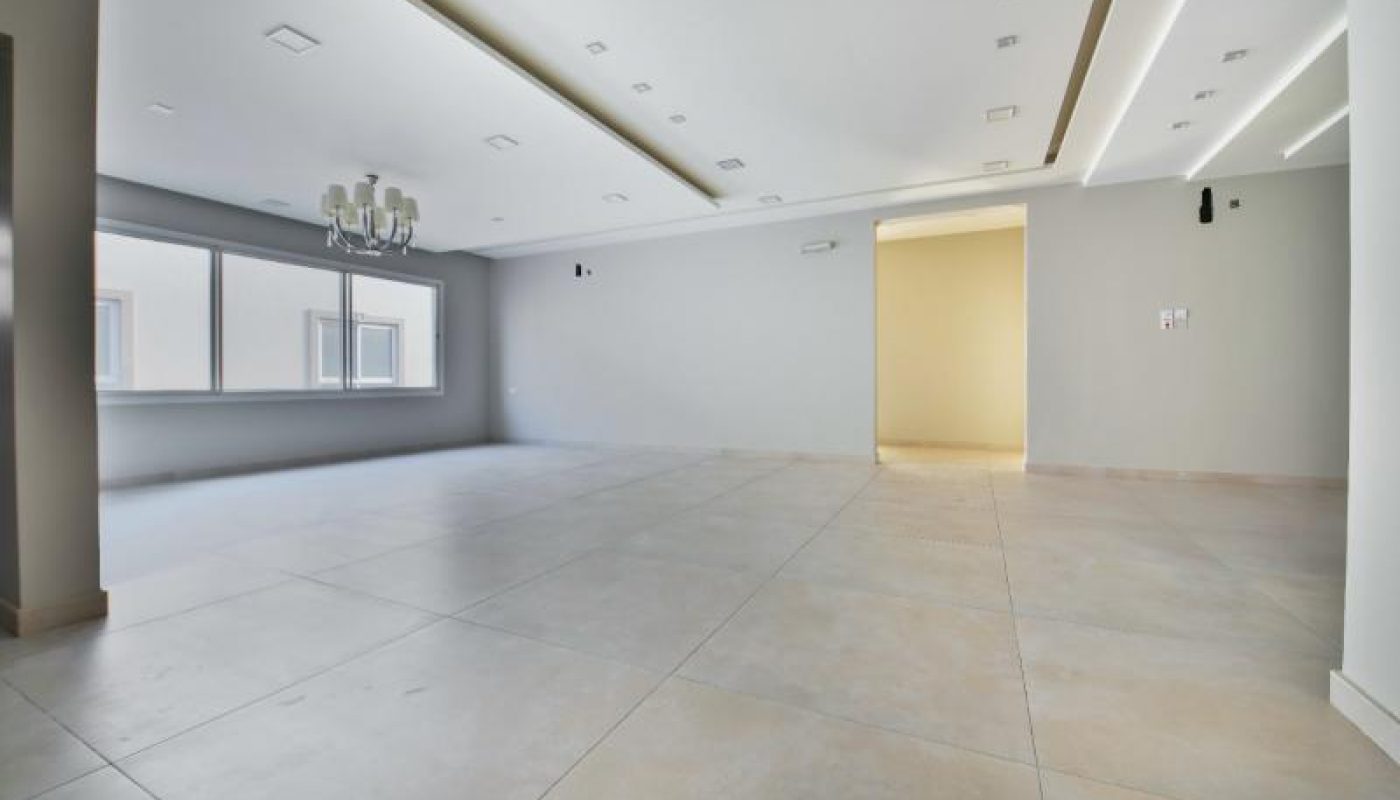 Spacious, empty room with large tiled floor, recessed ceiling lights, a chandelier, and large windows letting in natural light.