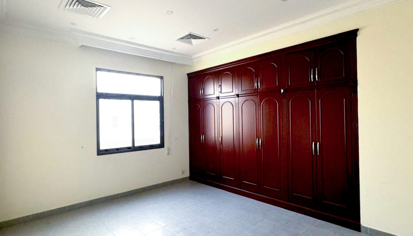 Empty room with tiled floor, cream-colored walls, a single window, and a large built-in wooden wardrobe with multiple doors. Ceiling has vents and recessed lights.