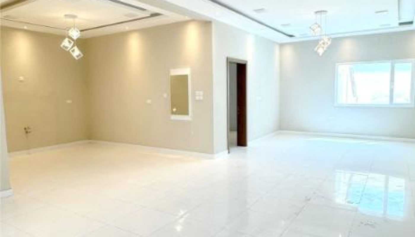 A spacious, empty room with white tiled floors, beige walls, recessed lighting, and a ceiling featuring multiple light fixtures and a window letting in natural light.