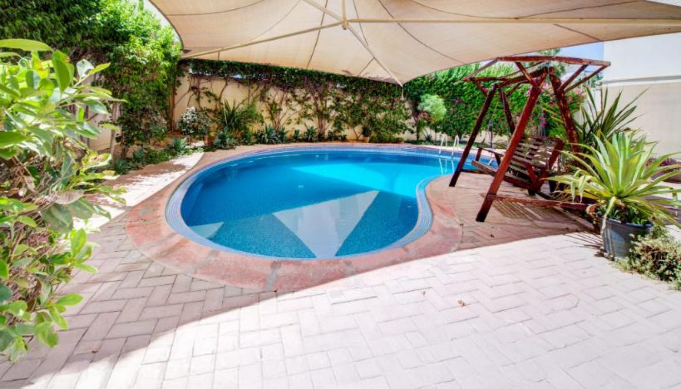 Small backyard pool with overhead shade canopy, surrounded by greenery, paved patio, and a wooden swing set on the right side.