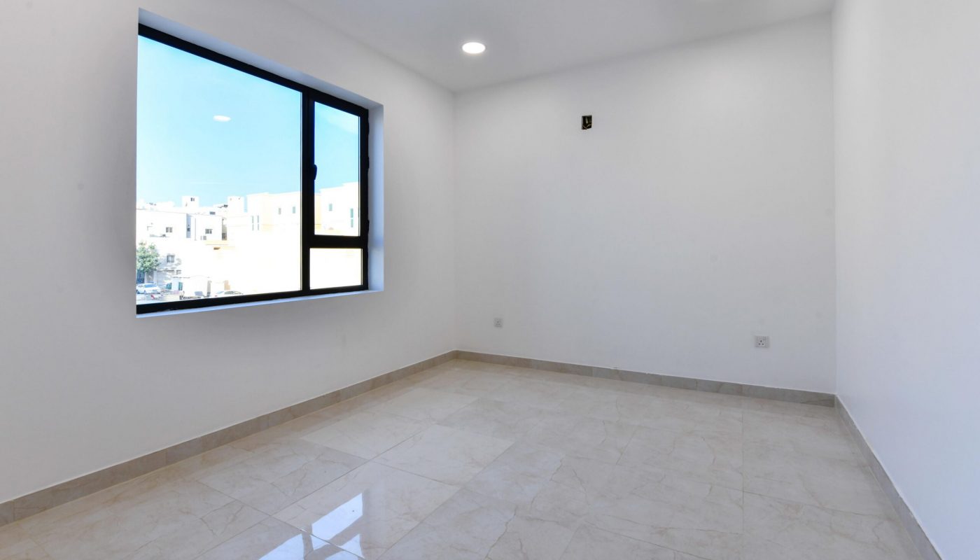 Empty, unfurnished room with white walls, large window, tiled floor, and recessed ceiling lights. Daylight is visible through the window.