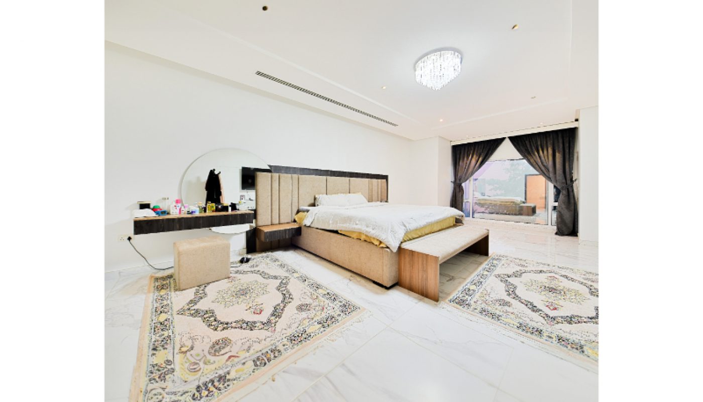 Spacious bedroom with a large bed, two patterned rugs, a vanity with a mirror, and sliding glass doors leading to a balcony with outdoor seating.