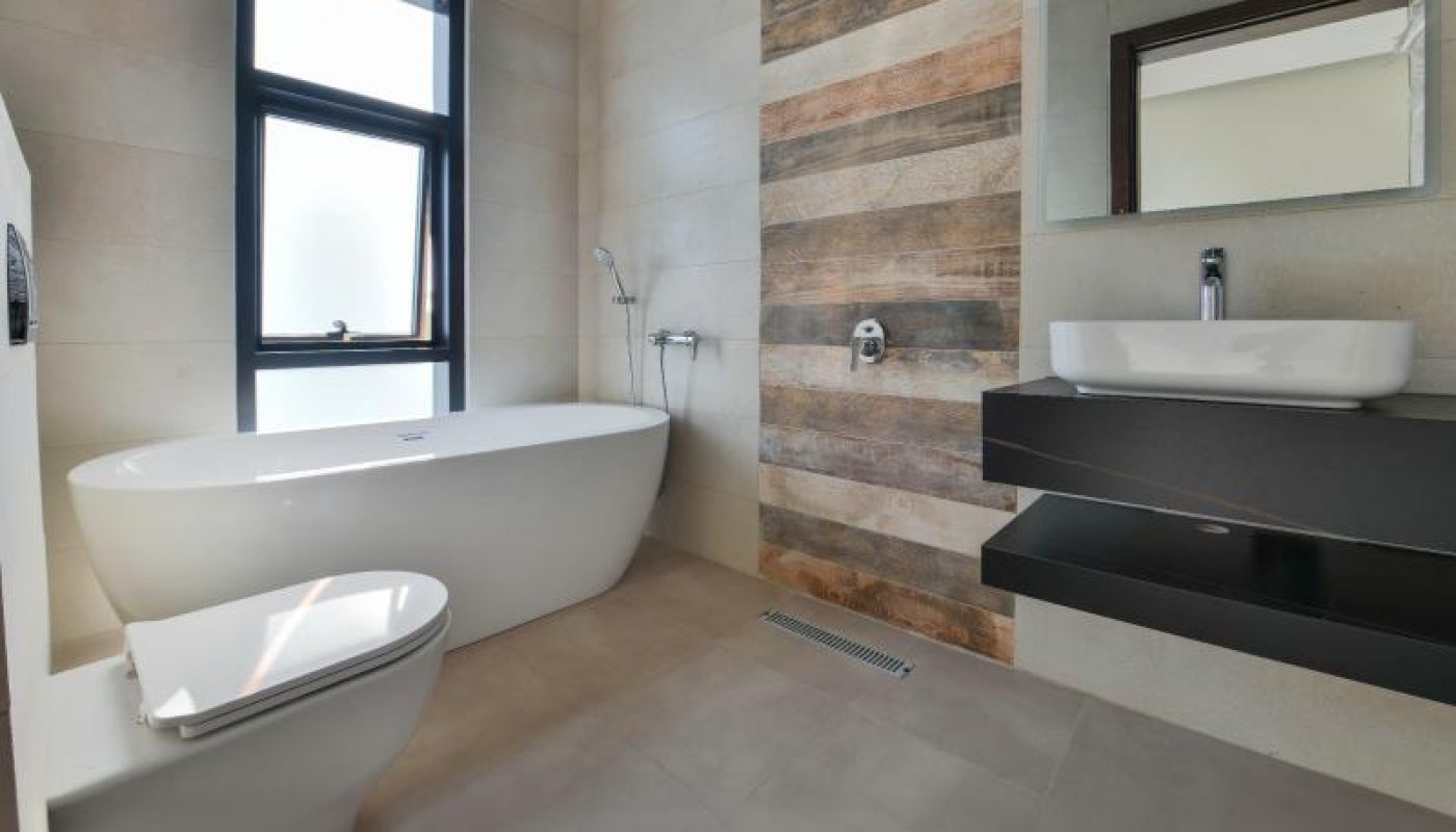 Modern bathroom with a freestanding bathtub, wall-mounted sink and mirror, toilet, large window, and wood-accented shower area.