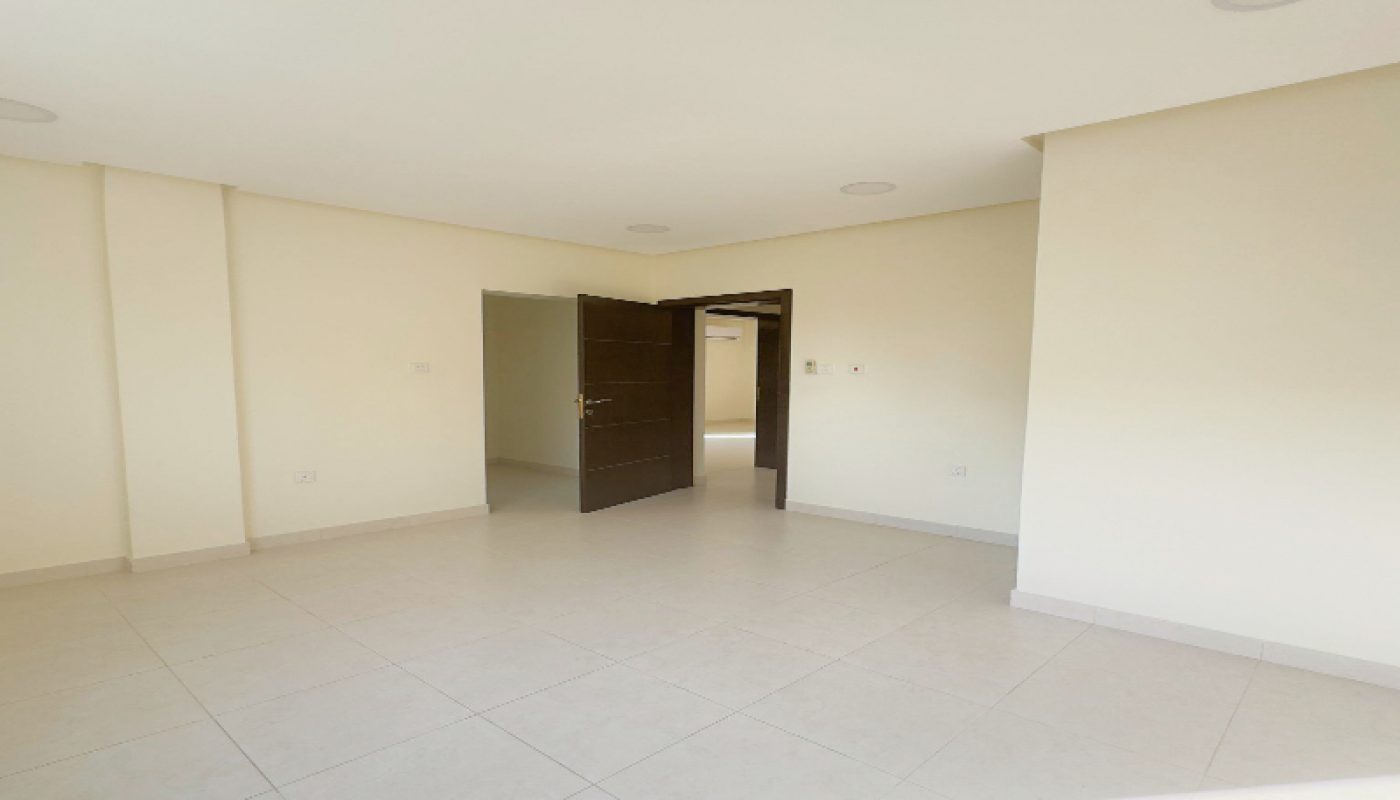 Empty, unfurnished room with white tiled floors, cream-colored walls, and an open dark wooden door leading to a hallway. Sunlight is entering from the right side of the image.