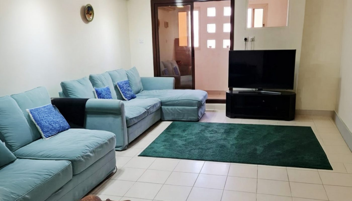 Living room with light blue sofas, green rug, flat-screen TV on a stand, wooden accent tables, tiled floor, and ceiling fan.