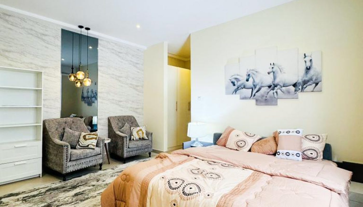 A modern bedroom with a double bed, two armchairs, a white shelving unit, and a wall art featuring white horses. Neutral tones and contemporary lighting complete the decor.