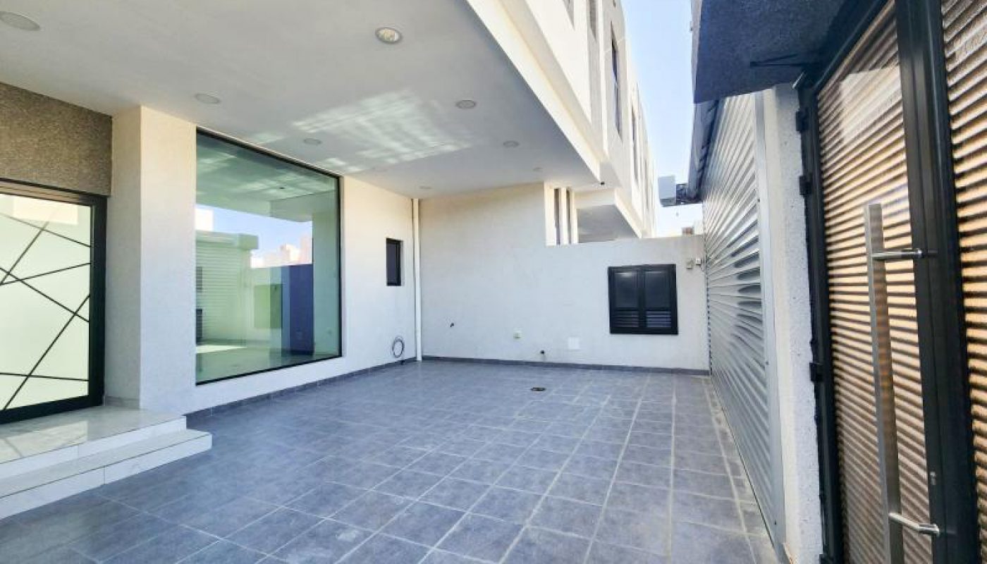 Modern outdoor patio with tiled flooring, large windows, white walls, and a metal gate on the right. Steps lead to a glass door; the area appears clean and spacious.